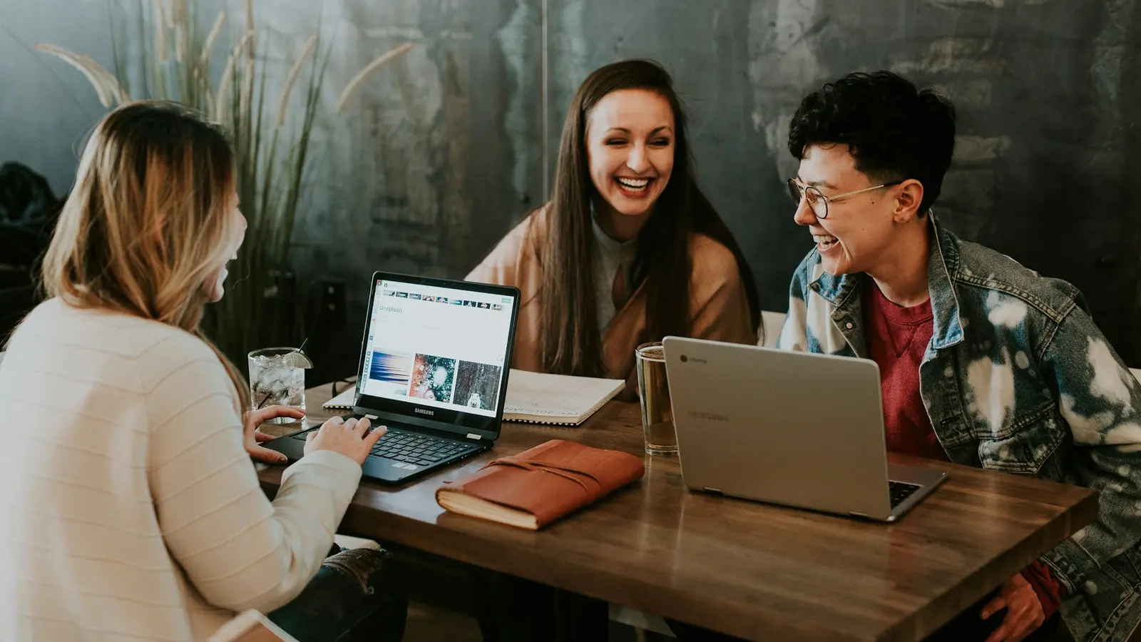 Drei Studierende lachen gemeinsam an einem Tisch mit Laptops und Notizbüchern