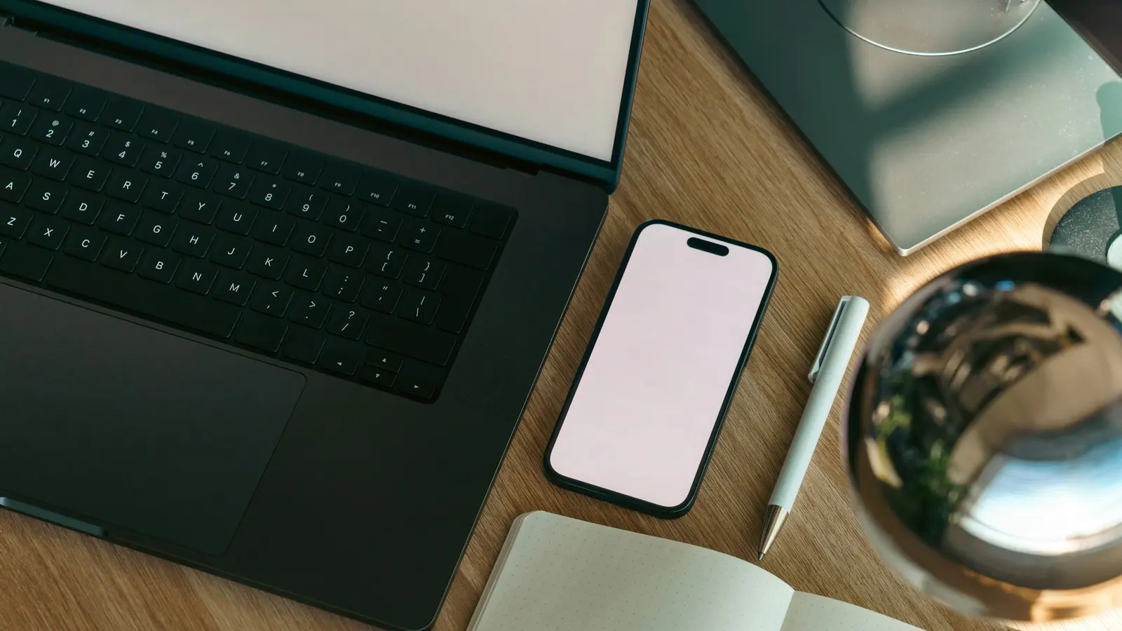 A laptop, a smartphone and a notebook on a wooden desk in daylight