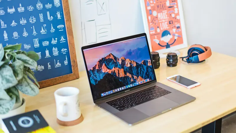 MacBook Pro sobre uma mesa de madeira ao lado de uma caneca branca, um celular e pôsteres coloridos