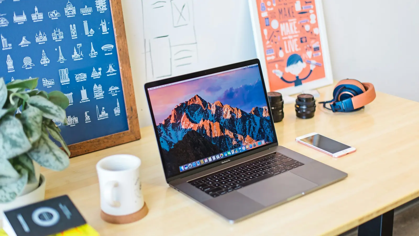 木桌上的MacBook Pro,旁边是白色马克杯、手机和色彩鲜艳的海报