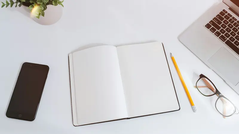 An open empty notebook on a white desk next to a smartphone and a laptop